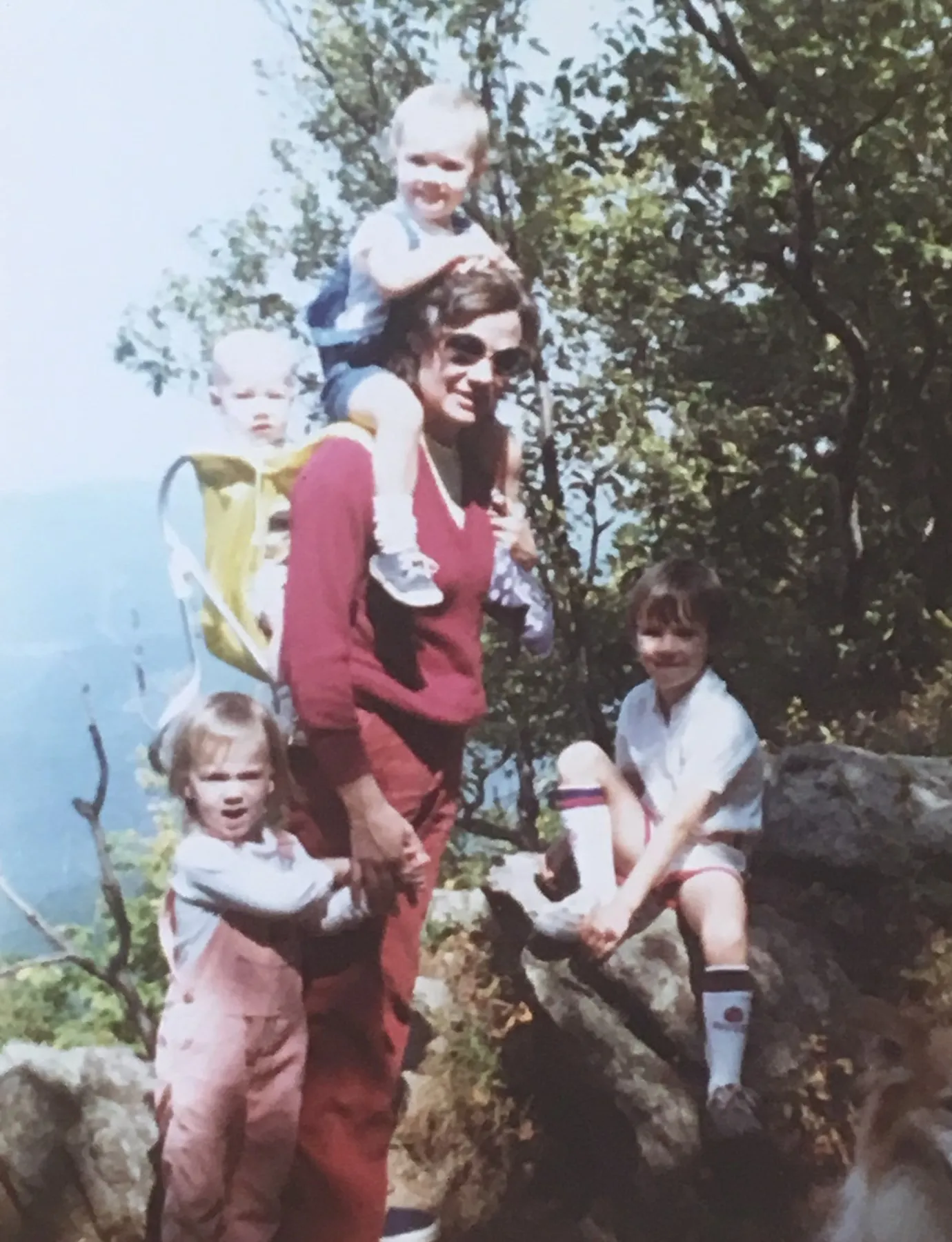 A young Christie on a mountain trail carrying one child on her shoulders and another in a backpack, with two more children beside her