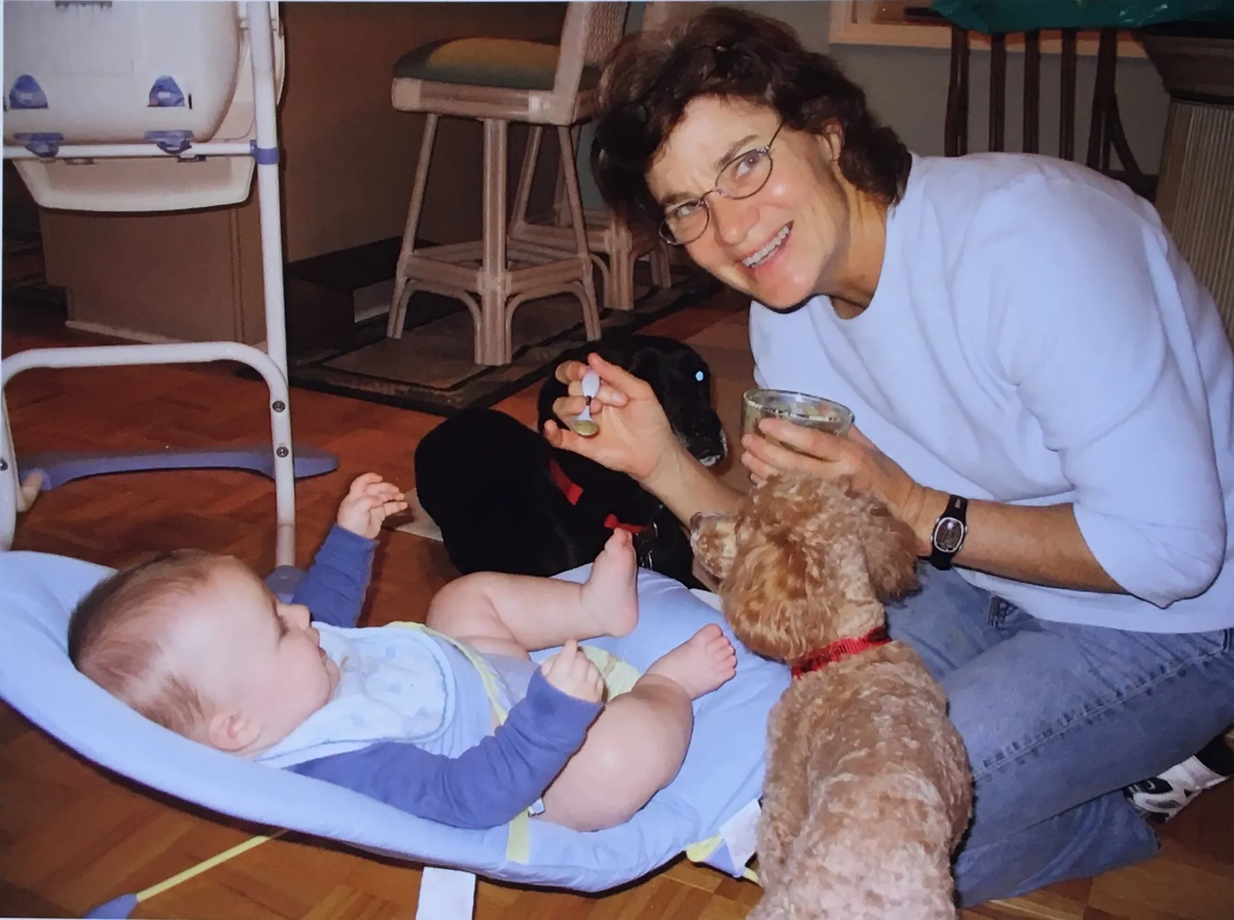 Christie kneeling on the floor, smiling, feeding a baby in a bouncer with two dogs beside her